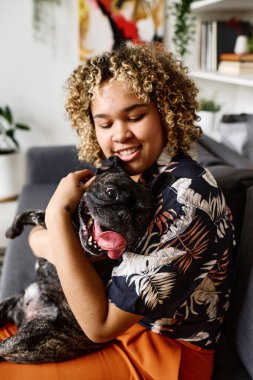 Kanepede oturup köpeğini kollarına alan kıvırcık saçlı Afrikalı genç kız evcil hayvanıyla bağ kuruyor.