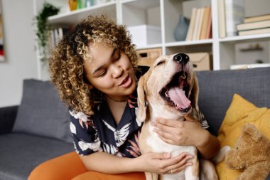 Kıvırcık saçlı Afrikalı genç kız oturma odasında kanepede otururken esnemiş köpeğiyle sarılıp konuşuyor.