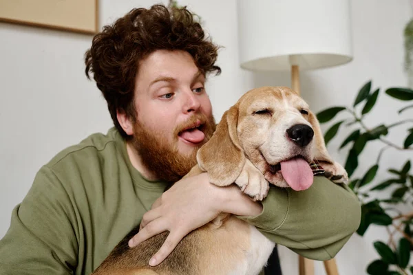 Young bearded man holding dog in his arms and sticking out his tongue just like his dog