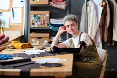 Genç, serbest çalışan, kısmi kollu kadın moda tasarımcısı. Masanın başında oturmuş, kağıt ve tekstil işleriyle uğraşırken yeni fikirler düşünüyor.