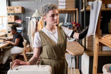 Moda koleksiyonu için yeni kıyafet modelleri seçerken kısmi kollu genç bir terzi kağıt üzerinde çizime bakıyor.