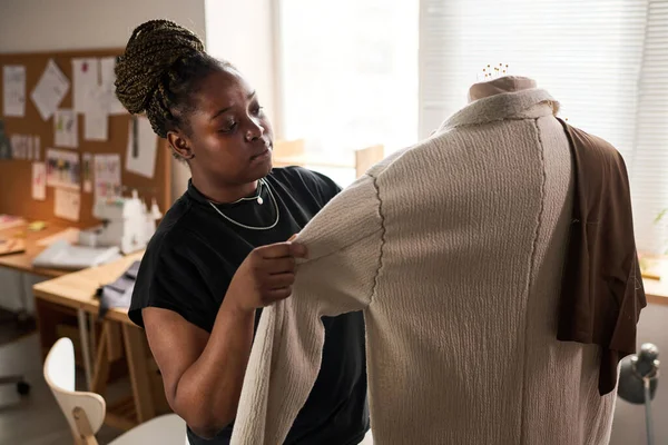 Young black woman looking at beige coat hanging on mannequin while creating new items for seasonal fashion collection in workshop