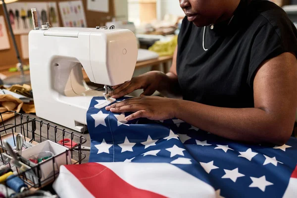 Young female volunteer sitting by electric sewing machine and making large American flag while preparing for Labor day