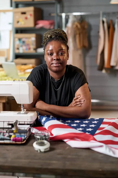 Young African American female tailor looking at camera while sitting by her workplace in front of electric sewing machine and USA flag