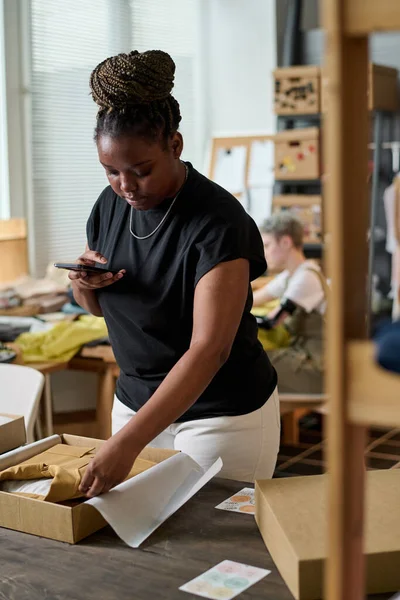 Young serious black woman with smartphone bending over workplace while taking photo of folded and packed clothes in workshop