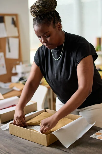 Young African American female worker of warehouse folding and packing new clothes in box before sending goods to receiver