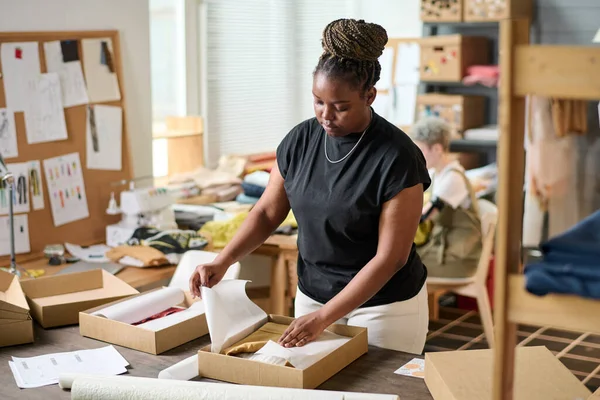 African American woman in casualwear standing by table and packing new clothes in cardboard box while wrapping goods into paper