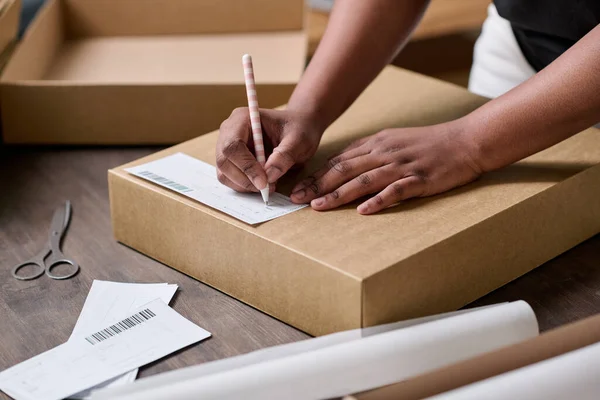 stock image Hands of young black woman with pen writing down address of client on top of cardboard box cover before packing ordered goods