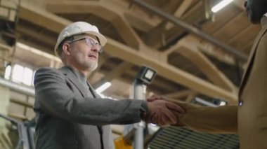 Beyaz şapkalı ve resmi kıyafetli kıdemli beyaz mühendisin el sıkışırken ve fabrikada Afrikalı Amerikalı meslektaşıyla konuşurken düşük açılı çekimi.