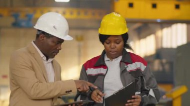 Afrika kökenli Amerikalı mühendis ve Afrikalı Amerikalı kadın teknisyen makine fabrikasında çalışırken dijital tablet ve pano üzerinde tartışıyorlar.
