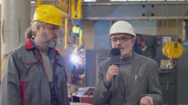 Kamerada mikrofonla konuşan bir TV muhabiri ve makine fabrikasında dururken kıdemli bir çalışanla röportaj yapan biri.