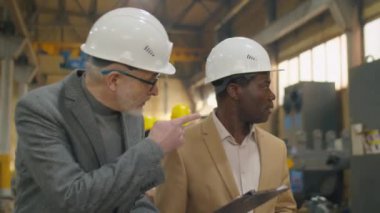 Kara şapka ve resmi giyimde kıdemli Kafkasyalı mühendis panoda bir şey açıklıyor ve Afrikalı Amerikalı meslektaşlarına endüstriyel fabrikada çalışırken makinelerden bahsediyorlar.