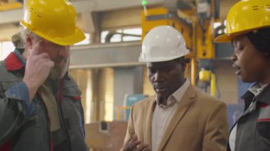 Siyah şapkalı ve resmi kıyafetli Afro-Amerikan mühendis makine fabrikasında iş birliği yaparken çeşitli erkek ve kadın teknisyenlerle dijital tablet üzerinde tartışıyor.