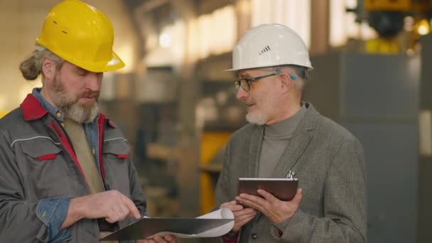 硬帽和正装高级工程师与成熟的男性技师一起在工厂工作时讨论数字平板和剪贴板上的一些问题 — 图库视频影像