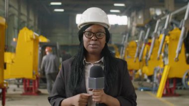 Afrika kökenli Amerikalı kadın muhabirin ağır ekipman fabrikasında dikilip kameraya bakarken ve mikrofonla konuşurken görüntüsü.