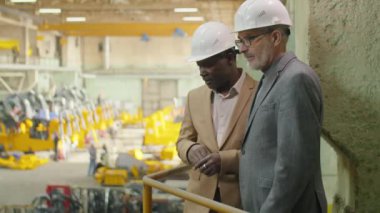 Orta yaşlı Afro-Amerikan ve Kafkasyalı mühendisler fabrikanın üst katında duruyor ve üretim sürecini tartışıyorlar.