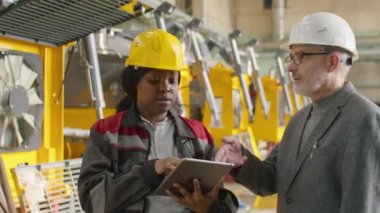 Afrika kökenli Amerikalı kadın mühendis dijital tablet kullanıyor ve makine fabrikasında çalışırken çok ırklı yöneticilerle konuşuyor.
