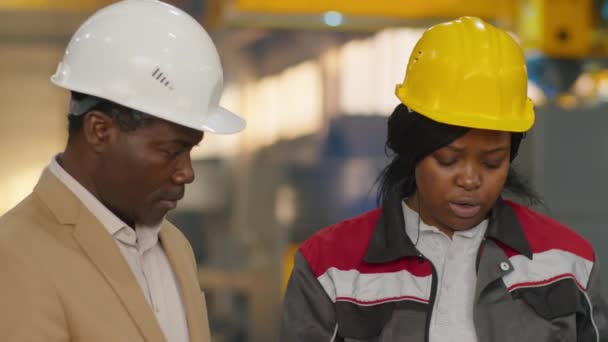 非洲裔美籍硬帽和正装工程师在机械厂一起工作时与非洲裔美籍女技师讨论纸制计划 — 图库视频影像