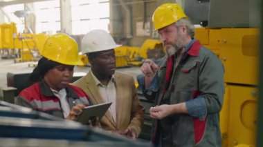 Dijital tablet kullanan ve fabrikada kalite kontrolü yaparken çok ırklı erkek meslektaşlarıyla konuşan Afro-Amerikan kadın.