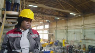 Üniformalı ve kasklı Afro-Amerikan kadın işçi fabrikaya iniyor ve koruyucu eldivenler giyiyor.