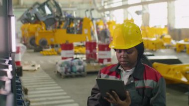 Afrika kökenli Amerikalı kadın kalite kontrol müfettişi fabrika deposundaki raflarda kutuları işaretliyor ve dijital tablet üzerinde çalışıyor.
