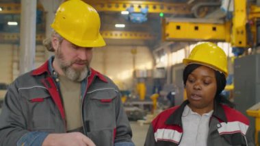Kıdemli Kafkasyalı erkek ve genç Afrikalı Amerikalı kadın kasketli ve iş kıyafetli birlikte fabrikada yürüyorlar, panoda kağıt okuyorlar ve makineleri tartışıyorlar.