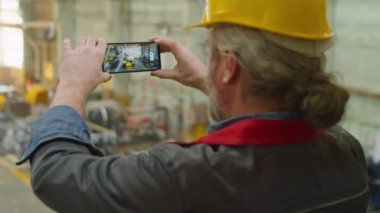 Fabrika kalite kontrol müfettişi. İş kıyafetleri ve kasketi. Akıllı telefonlu makinelerin fotoğrafını çekiyor.