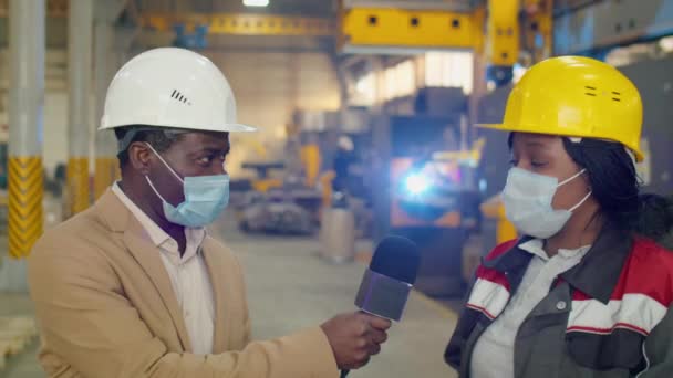アフリカ系アメリカ人のテレビ記者は ハードハットとマイクで話す保護マスクで 流行病の流行中に女性工場技術者にインタビューしながら — ストック動画