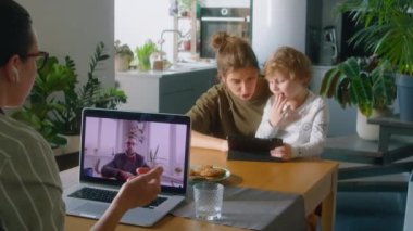 Karısı ve küçük oğlu evdeki mutfak masasında dijital tablet kullanırken, iş kadını kablosuz kulaklıklarla iş arkadaşıyla bilgisayardan görüntülü konuşma yapıyor.