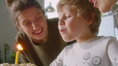 Mutlu küçük çocuk doğum günü pastasında mum üflerken neşeli lezbiyen anneler evde onu öpüyor ve gülümsüyor.