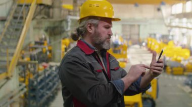 Fabrikada akıllı telefon kullanan kasklı ve üniformalı kıdemli sanayi işçisi.