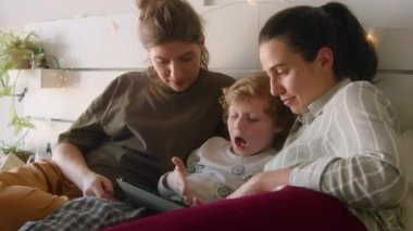 Küçük çocuk evde yatakta yatarken lezbiyen annelerle dijital tablette bir şeyler konuşuyor.