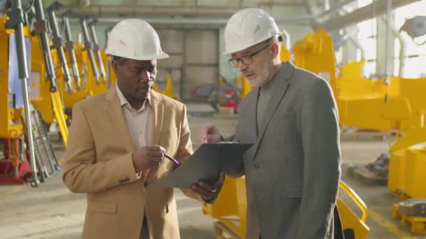 成熟的白种人和非洲裔美国男性经理 头戴硬帽 身穿正装 在机械厂一起工作时 在剪贴板上讨论一些事情 — 图库视频影像