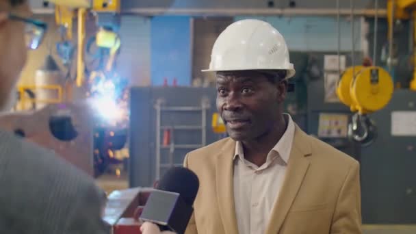 テレビジャーナリストに工場についてのインタビューをしながら ハードハットでアフリカ系アメリカ人男性エンジニアの肩のショットとマイクで話す正式なスーツの上に — ストック動画