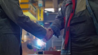 Resmi kıyafetli bir mühendis ve iş elbiseli bir teknisyen el sıkışıyor ve fabrikada iş yerinde tartışıyorlar.