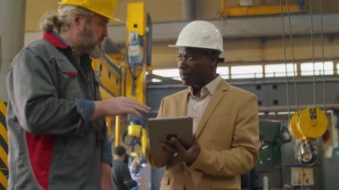Dijital tablet kullanan ve makine fabrikasındaki Kafkasyalı üst düzey teknisyenle iş tartışan sıkı şapka ve resmi kıyafet giymiş olgun Afrikalı Amerikalı mühendis.