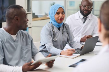 Genç tesettürlü kadın doktor, ofiste konferans sırasında dizüstü bilgisayar kullanıyor ve iş arkadaşlarıyla konuşuyor.
