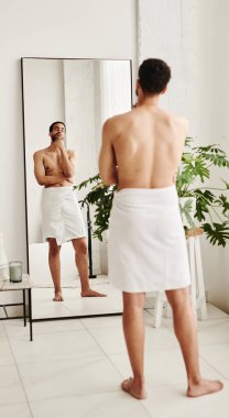 Havlu giymiş genç bir adam aynaya bakıyor ve duştan sonra kendisine hayran.