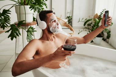 Yüz maskeli genç adam şarap içiyor ve köpükle banyo yaparken cep telefonunda selfie portresi yapıyor.