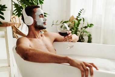 Genç adam banyoda dinlenmenin tadını çıkarıyor, kablosuz kulaklıkla müzik dinliyor ve kırmızı şarabın tadını çıkarıyor.