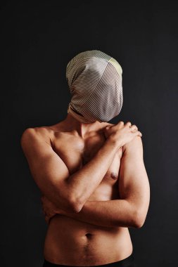 Üstsüz, kaslı bir adamın portresi. Kendini kucaklıyor. Siyah arka planda, başında eşarpla dikiliyor.
