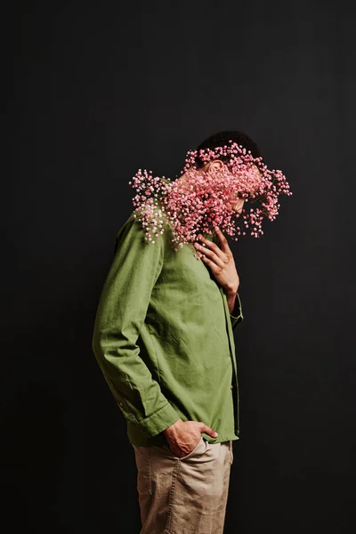 Side view of young man in green shirt hiding behind the pink dried flowers against the black background