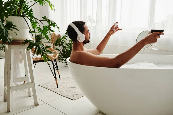 在浴室的温泉疗程中 年轻人一边喝酒一边在浴缸里放松 一边用耳机欣赏音乐 — 图库照片