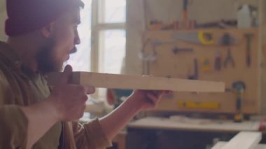 Erkek marangoz ahşap kalas tutuyor ve marangozluk atölyesinde ondan toz üflüyor.