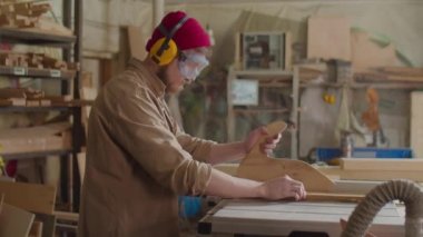 Güvenlik kulaklığı ve gözlüklü bir erkek marangoz, ahşap atölyesinde tahta kesmek için masa testeresi kullanıyor.