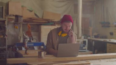 Boynunda güvenlik kulaklıkları olan, cep telefonuyla konuşan ve marangozluk atölyesinde dizüstü bilgisayar kullanan beyaz erkek ahşap işçisi.