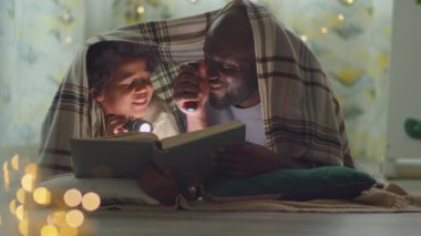 Afrikalı Amerikalı baba ve küçük oğlu battaniyenin altında yatıyorlar, ellerinde fenerler, kitap okuyor ve akşam evde sohbet ediyorlar.