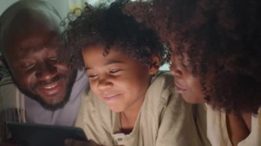Afrika kökenli Amerikalı anne, baba ve küçük oğlu karanlık bir odada birlikte yatıyorlar, gülümsüyorlar ve dijital tabletlerden bir şeyler izliyorlar.