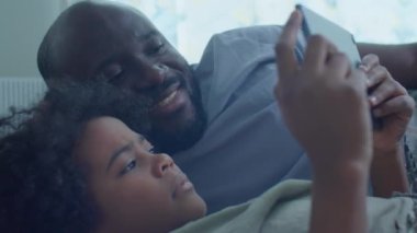 Afro-Amerikalı baba ve küçük oğlu evde boş zamanlarını geçirirken dijital tablette birlikte yatıp video izliyorlar.