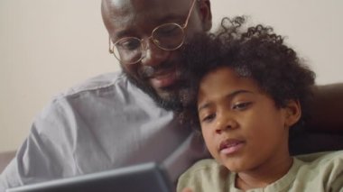 Neşeli Afrikalı Amerikalı baba ve küçük oğlu evdeki kanepede oturup dijital tablette bir şeyler izleyip konuşuyorlar.